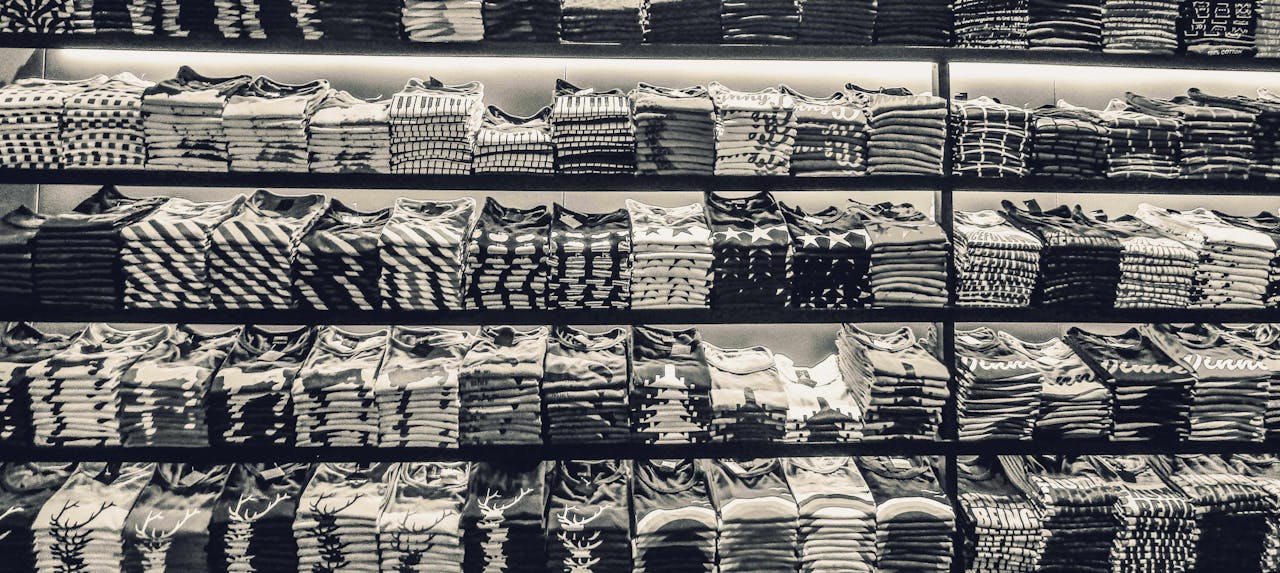 about-us-02 Black and white image showcasing neatly folded t-shirts stacked on store shelves.